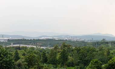 Yoğun orman ve dağ sıralarıyla çevrili uzak sanayi binaları bulutlu bir gökyüzü altında, doğa ve gelişim arasındaki çevresel zıtlığı gösteriyor..