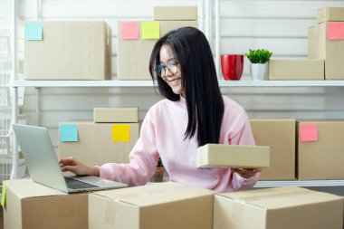Gülümseyen kadın bir dizüstü bilgisayar üzerinde çalışırken çevrimiçi mağazası için paketler hazırlıyor, e-ticaret iş akışını, küçük işletme faaliyetlerini ve tatmin faaliyetlerini gösteriyor.