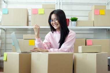 Sevkiyat kutuları ve dizüstü bilgisayarla çevrimiçi mağazasını yöneten gülümseyen kadın e-ticaret başarısını, küçük iş akışını ve modern girişimciliği ifade ediyor..