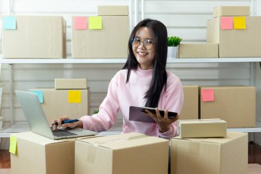 Sevkiyat kutuları arasında dizüstü bilgisayar ve tablet kullanarak çevrimiçi mağaza siparişlerini yöneten, e-ticaret iş akışını, envanter organizasyonunu ve küçük işletme operasyonlarını gösteren gülümseyen kadın..