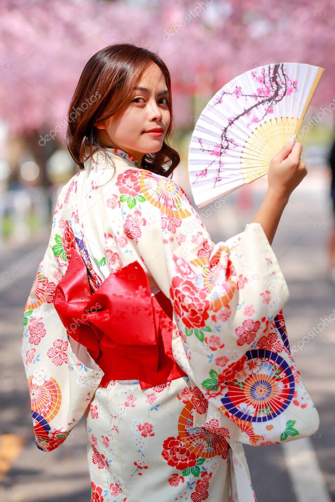 Portrait of a traveler young asian woman wear a dress yukata and walking on tree path in japan cherry blossoming sakura park at sunlight afternoon.