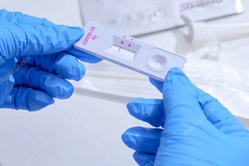Woman wearing blue medical gloves and holding COVID-19 (SARS-CoV-2) First Antigen Test kit device to help in the Rapid Detection testing at home. Showing negative COVID-19 outbreak.