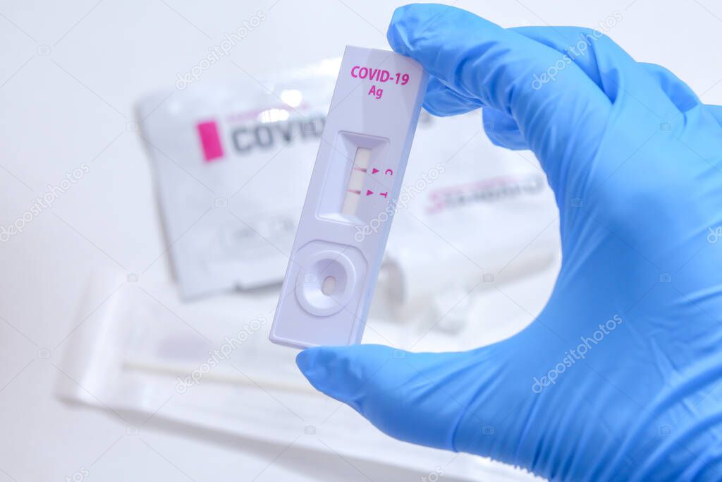 Woman wearing blue medical gloves and holding COVID-19 (SARS-CoV-2) First Antigen Test kit device to help in the Rapid Detection testing at home. Showing positive COVID-19 outbreak.