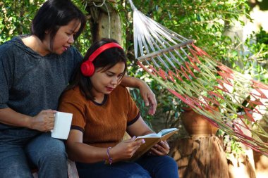 Asyalı bir kadın bahçede oturuyor, kitap okuyan bir kız ve diğeri kolunu omzuna koyup elinde kahve fincanı tutuyor..