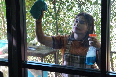 Asyalı bir kadının portresi temizleyici spreyliyor ve pencereyi siliyor. İfadesine ve temizlik faaliyetlerine odaklanıyor.