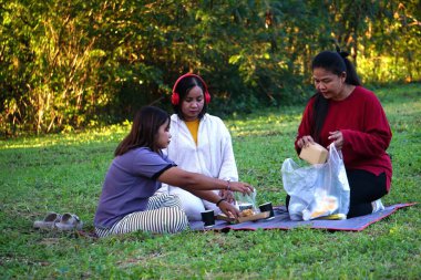 Üç kadın dinleniyor ve piknik minderinde sohbet ediyorlar. Akşam güneşinin yumuşak ışığıyla aydınlatılıyorlar..