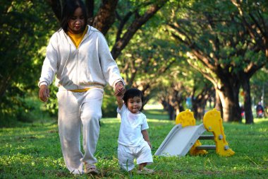 Asyalı bir kadın küçük bir çocuğu kucağında tutuyor ve parkta çocuğa sevgiyle gülümsüyor.