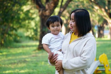 Bir kadın küçük bir çocuğu kucağında tutuyor ve parkta çocuğa sevgiyle gülümsüyor.