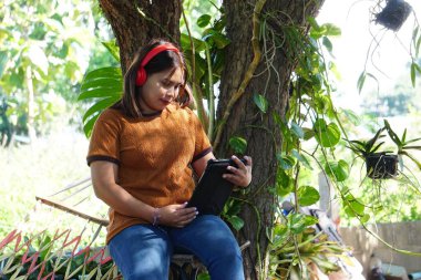 Kahverengi bluzlu Asyalı kadın bir ağacın altında oturmuş tablete bakıyor..