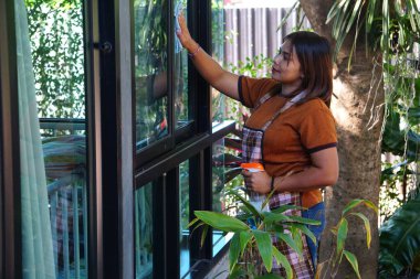 Asyalı kadın bir elinde bez, diğerinde sprey şişeyle uzun bir pencereyi temizlerken hafifçe yukarı bakar..