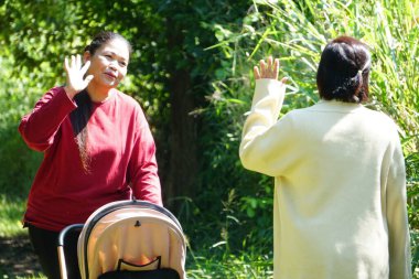 İki kadın bir bebek arabasının yanında karşı karşıya durur ve yemyeşil bir alanda ellerini sallarlar..