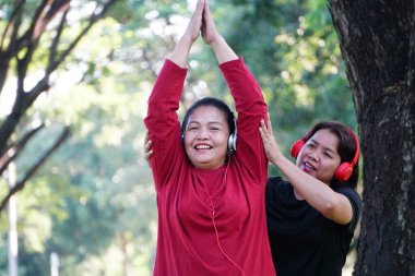 Kırmızı gömlekli ve kulaklıklı bir kadın oturuyor ve kollarını başının üstüne kaldırmış, arkasında siyah elbiseli başka bir kadınla esniyor..