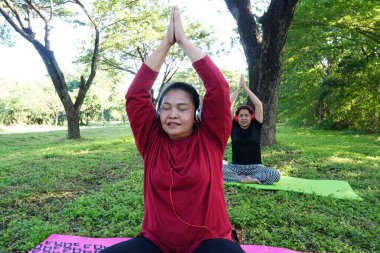 Kırmızı gömlekli ve kulaklıklı bir kadın oturuyor ve kollarını başının üstüne kaldırmış, arkasında siyah elbiseli başka bir kadınla esniyor..