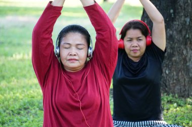 Kırmızı gömlekli ve kulaklıklı bir kadın oturuyor ve kollarını başının üstüne kaldırmış, arkasında siyah elbiseli başka bir kadınla esniyor..