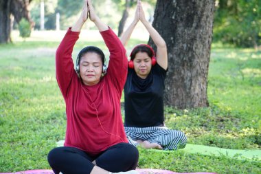 Kırmızı gömlekli ve kulaklıklı bir kadın oturuyor ve kollarını başının üstüne kaldırmış, arkasında siyah elbiseli başka bir kadınla esniyor..