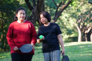 İki kadın parkta dikilip çene çalıyorlar, ellerinde picleball raketleri ve yeşil bir top..
