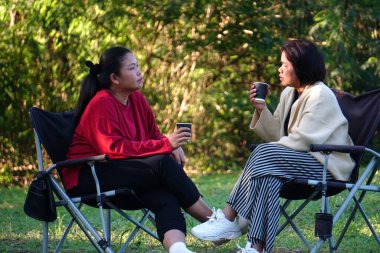 Asyalı iki kadın sandalyede oturmuş açık havada içki içiyor..