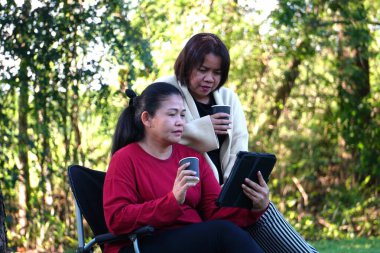 İki kadın dışarıda ayakta ve dinleniyor, biri elinde kahve fincanı, diğeri de arkadaşının arkasında durduğu tableti izliyor..