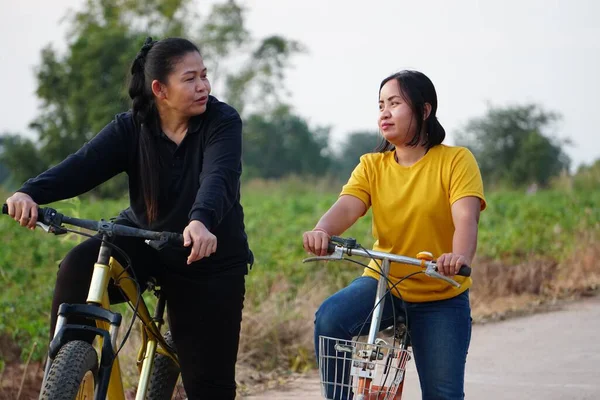Asyalı kadınlar kırsal bir yolda bisiklet sürmeye ara verir ve birbirleriyle yakın konuşurlar..