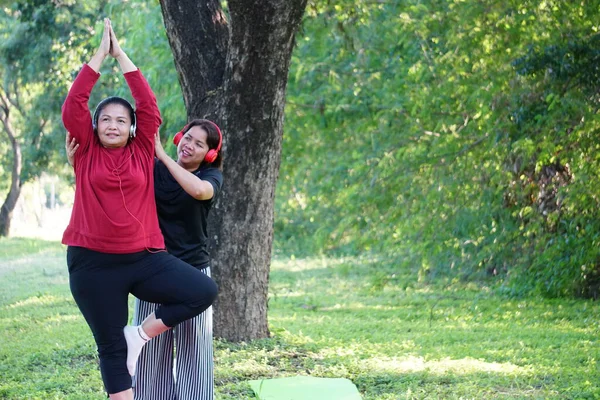 İki kadın mutlu bir şekilde gülümserken, bir kadın (siyah gömlekli) kollarını kaldırmış yoga pozu veren kırmızı gömlekli arkadaşına yardım ediyor..
