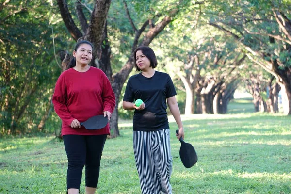 İki kadın parkta dikilip çene çalıyorlar, ellerinde picleball raketleri ve yeşil bir top..