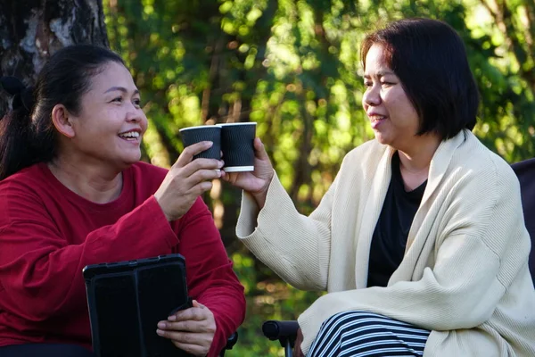 İki kadın dışarıda ayakta ve dinleniyor, biri elinde kahve fincanı, diğeri de arkadaşının arkasında durduğu tableti izliyor..