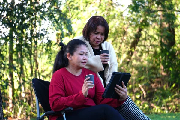 İki kadın dışarıda ayakta ve dinleniyor, biri elinde kahve fincanı, diğeri de arkadaşının arkasında durduğu tableti izliyor..