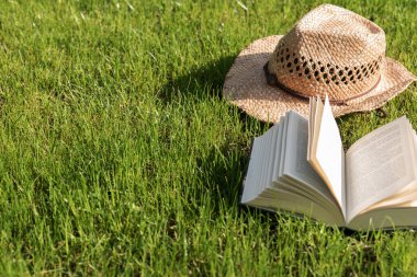 Açık bir kitap arka planda hasır şapkayla çimenlikte yatıyor. Koronavirüs salgını sırasında bahçede rahatlama