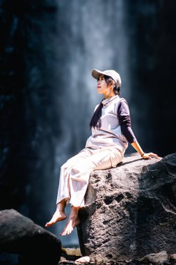 Arka planda Dolo şelalesi olan büyük bir kayanın üzerinde oturan Asyalı bir kadın. Doğada tatilin tadını çıkarıyorum..