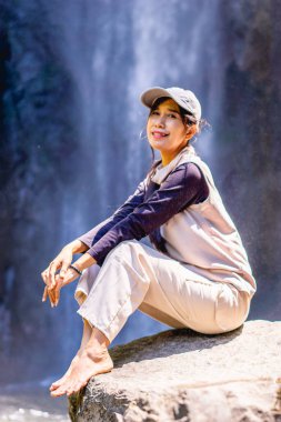 Arka planda Dolo şelalesi olan büyük bir kayanın üzerinde oturan Asyalı bir kadın. Doğada tatilin tadını çıkarıyorum..
