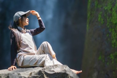 Arka planda Dolo şelalesi olan büyük bir kayanın üzerinde oturan Asyalı bir kadın. Doğada tatilin tadını çıkarıyorum..