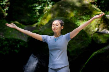 Doğada yoganın keyfini çıkaran Asyalı bir kadın. Büyük yosun kayaları ve ormanda akan nehir suyuyla.