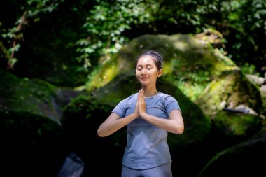Doğada yoganın keyfini çıkaran Asyalı bir kadın. Büyük yosun kayaları ve ormanda akan nehir suyuyla.