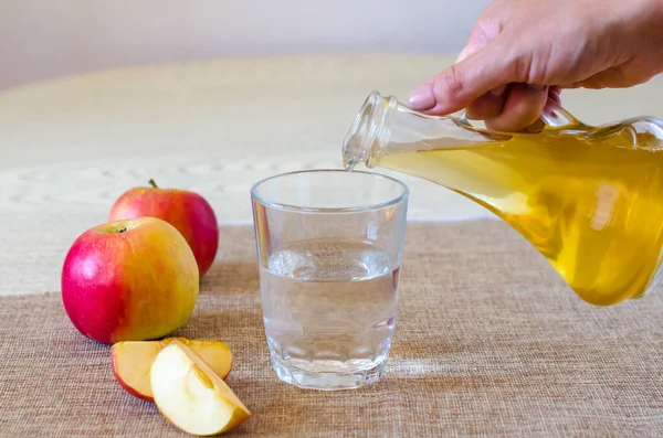 Sebuah tangan wanita menuangkan cuka sari apel dalam botol kaca pada latar belakang cahaya ke dalam segelas air. Asam malat bermanfaat bagi kesehatan dan digunakan dalam memasak. — Stok Foto