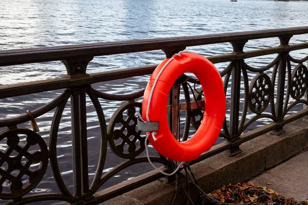 Köprü korkuluğu ve üzerinde turuncu bir can simidi olan bir nehir manzarası. Kompozisyon, su kenarında sakin bir şehir anını yakalıyor, güvenlik, renk kontrastı ve günlük basit ayrıntıları vurguluyor..
