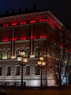 Riga 'daki tarihi bir bina, geceleri çarpıcı kırmızı ve sıcak beyaz ışıklarla aydınlatılıyor. Süslü sokak lambaları ve sonbahar ağaçlarıyla vurgulanarak dramatik ve atmosferik bir şehir manzarası yaratıyor..