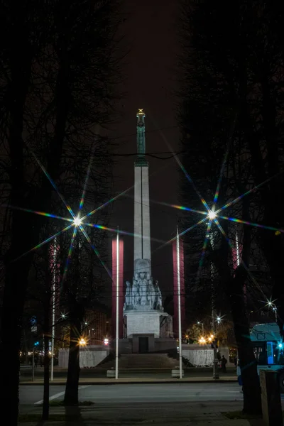 A night-time view of the Freedom Monument in Riga, Latvia, illuminated against the dark sky. The monument stands tall between rows of bare trees, with bright starburst streetlights and Latvian flags on both sides. 