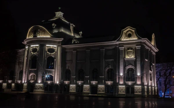 An elegant historic building in Riga illuminated at night, showcasing ornate architectural details and soft lighting that highlights its grand faade against the dark sky.