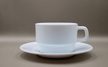 Tarafsız bir arkaplanda fincan tabağı olan minimalist beyaz seramik kahve fincanı. Yumuşak ışıklandırma ve kopyalama alanı olan temiz ve basit kompozisyon. Kahve kavramları, kafe markalaşması, yaşam tarzı tasarımı, sabah rutinleri, sakin anlar ve modern minimal tasarım için idealdir..