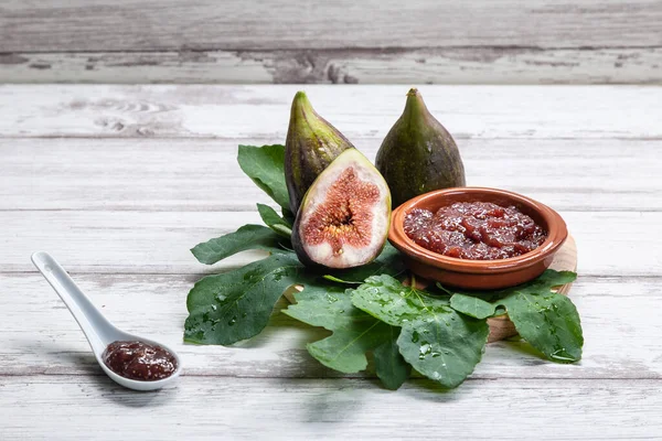 Fig jam with figs, fig leaves and white spoon with jam on white wooden background.