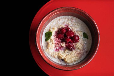 Kiraz ve nane yapraklarıyla süslenmiş kinoa lapası. Güzel bir seramik kırmızı fincanda kırmızı bir masanın üstünde duruyor, üst manzara. Sağlıklı ve sağlıklı bir kahvaltı..