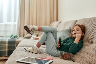 Televizyon izleyen küçük tatlı bir kız elinde uzaktan kumandayla kanepede yan yana oturuyor.