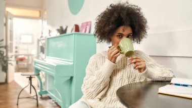 Hoş, koyu tenli bir kadın şık kıyafetler giyip kahve içerken kameraya bakıyor ve bir sandalyede oturuyor.
