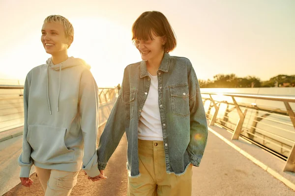 Ser amado. Feliz pareja de lesbianas riendo, tomados de la mano mientras caminan por el puente y observan el amanecer juntos —  Fotos de Stock