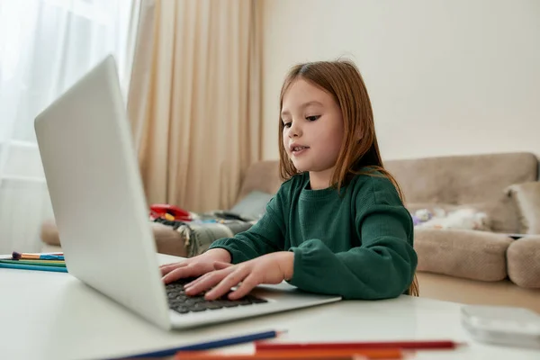 Uma menina pequena bonito gosta de conversar com seus amigos através de um laptop enquanto sentado sozinho em um enorme quarto brilhante sozinho durante a educação à distância — Fotografia de Stock