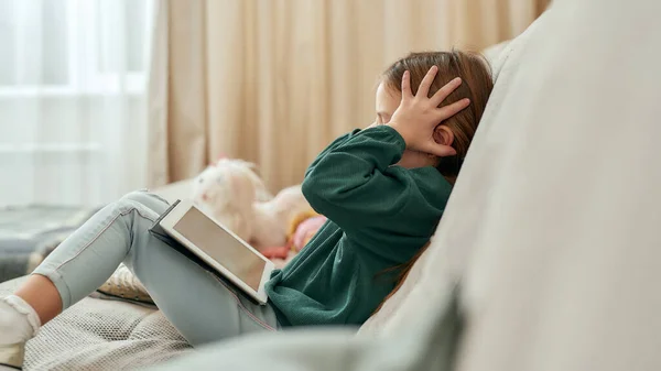 Uma garotinha fofa expressando emoções sobre um videogame em um tablet enquanto sentada em um sofá com as mãos nos ouvidos — Fotografia de Stock