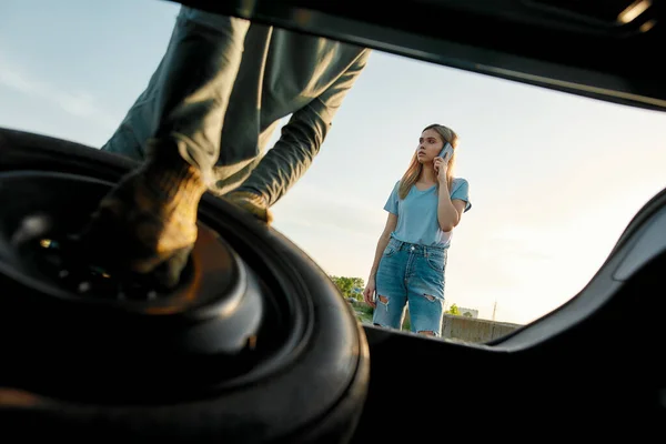 Young woman calling car service or assistance while having troubles with her car, Close up of hands of man taking out spare wheel — Stock Photo, Image