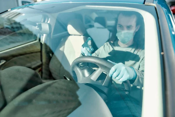 Man met medisch masker en beschermende handschoenen die antibacteriële desinfecterende spray op het stuur spuiten terwijl hij het interieur van de auto reinigt om virussen te voorkomen — Stockfoto
