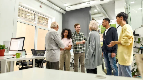 Glada unga laget hälsar på nya anställda, Åldrad man och kvinna, seniorpraktikanter skakar hand med kollegor på det moderna kontoret — Stockfoto