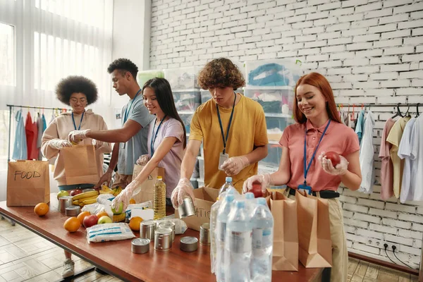 Young male and female volunteers packing food and drinks donation for homeless into paper bags, Small group of people working in charitable foundation — Stock Photo, Image